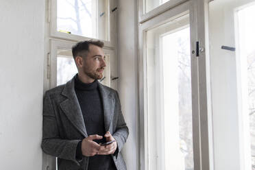 Junger Mann mit Mobiltelefon am Fenster stehend - GUSF02068