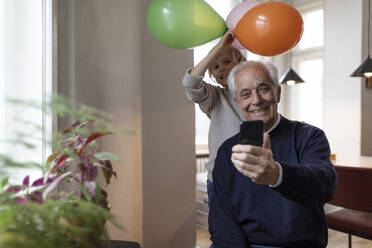 Happy grandfather and grandson taking a selfie with balloons at home - GUSF02085