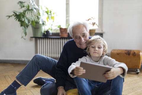 Großvater und Enkel sitzen zu Hause auf dem Boden und benutzen ein Tablet, lizenzfreies Stockfoto