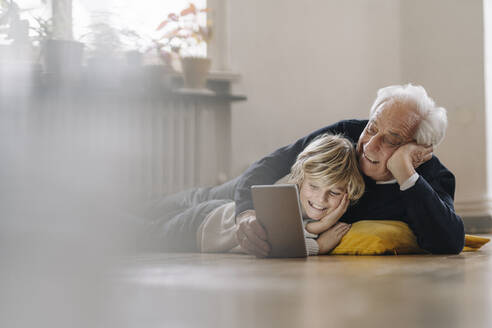 Happy grandfather and grandson lying on the floor at home using a tablet - GUSF02183
