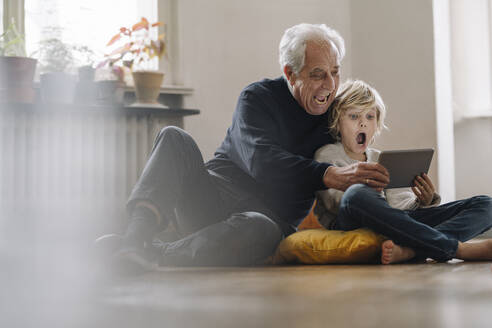 Screaming grandfather and grandson sitting on the floor at home using a tablet - GUSF02189