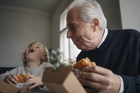 Happy grandfather and grandson eating burger together at home - GUSF02197