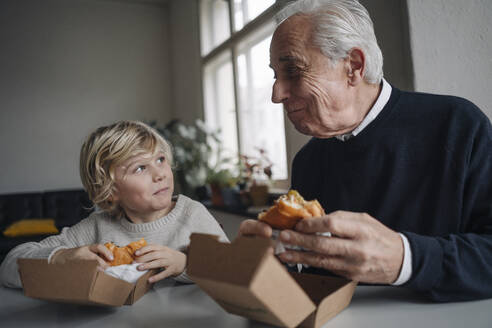 Smiling grandfather and grandson eating burger together at home - GUSF02198