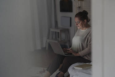 Ältere Frau sitzt zu Hause auf dem Bett und benutzt einen Laptop - GUSF02220