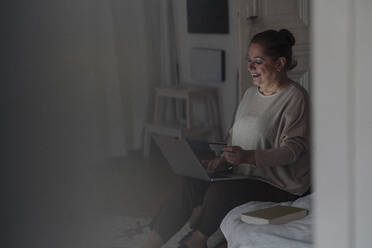 Happy senior woman sitting on bed at home using credit card and laptop - GUSF02221