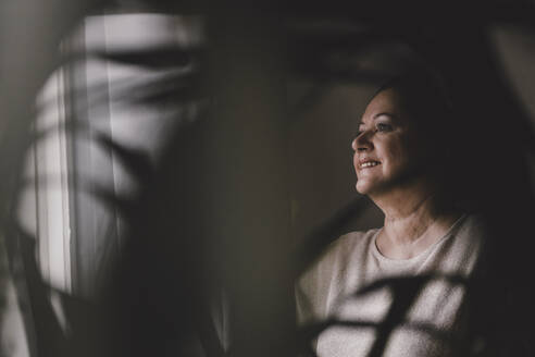 Smiling senior woman looking out of window at home - GUSF02223