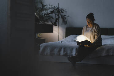 Senior woman sitting on bed holding glowing book - GUSF02225