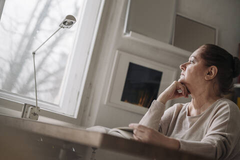 Ältere Frau mit Buch zu Hause und Blick aus dem Fenster, lizenzfreies Stockfoto