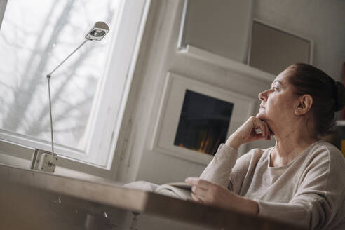Ältere Frau mit Buch zu Hause und Blick aus dem Fenster - GUSF02229