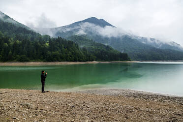 Mann steht am Seeufer und macht ein Foto mit seinem Smartphone, Wiestal-Stausee, Bundesland Salzburg, Österreich - JUNF01665