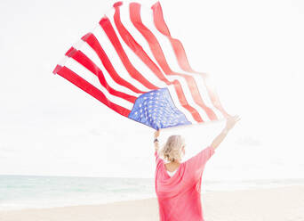 Ältere kaukasische Frau hält amerikanische Flagge am Strand - BLEF10433