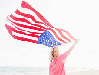 Ältere kaukasische Frau hält amerikanische Flagge am Strand - BLEF10434