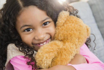 Mixed race girl hugging teddy bear on sofa - BLEF10463
