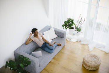 Smiling woman sitting on couch at home with laptop - SODF00028