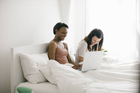 Zwei glückliche Frauen liegen zu Hause im Bett und benutzen Laptops, lizenzfreies Stockfoto