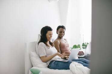 Zwei glückliche Frauen liegen zu Hause im Bett und benutzen Laptops - SODF00035