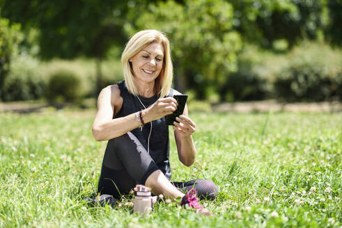 Ältere Frau benutzt ihr Smartphone und ruht sich nach dem Sport im Gras eines Parks aus - JSMF01148