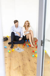 Happy couple in businesswear sitting on the floor surrounded by colourful balls - PESF01681