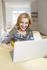 Laughing young woman using laptop on couch at home - PESF01711
