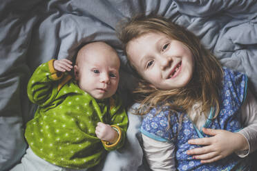 Portrait of smiling little girl lying on bed beside baby girl, top view - IHF00177