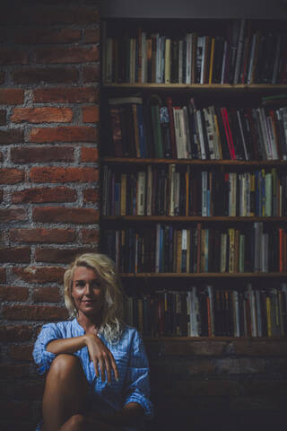 Lächelnde Frau sitzt in der Nähe eines Bücherregals, lizenzfreies Stockfoto