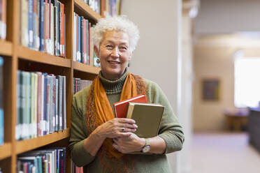 Ältere gemischtrassige Frau hält Bücher in einer Bibliothek - BLEF10831