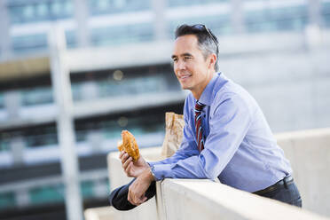 Gemischtrassiger Geschäftsmann isst auf dem Dach einer Stadt - BLEF10952