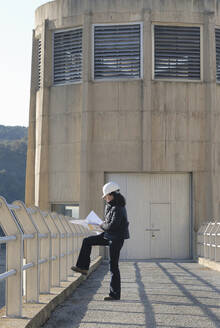 Weiblicher Ingenieur, der eine technische Zeichnung außerhalb eines Wasserkraftwerks betrachtet, Seitenansicht - BLEF11108