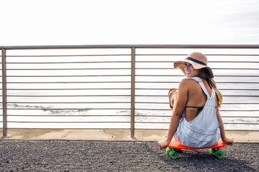 Kaukasische Frau sitzt auf einem Skateboard am Strand - BLEF11626