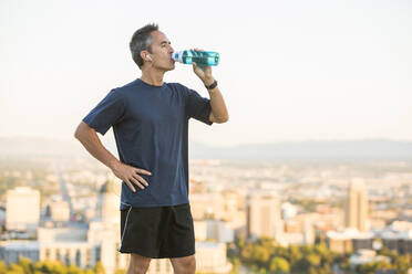 Mixed race man drinking water bottle on urban hilltop - BLEF11639