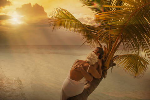 Older Caucasian couple hugging on palm tree at beach stock photo