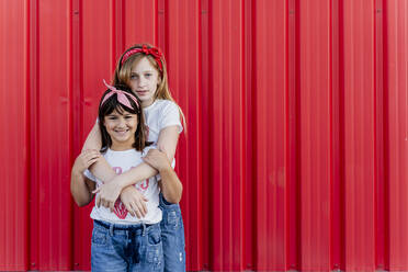 Sisters hugging in front of a red wall - ERRF01614