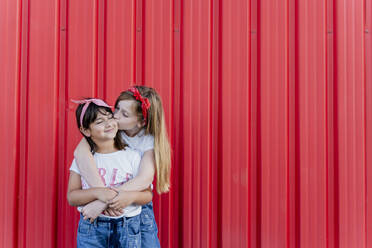 Two sisters standing in front of a red wall, kissing - ERRF01616