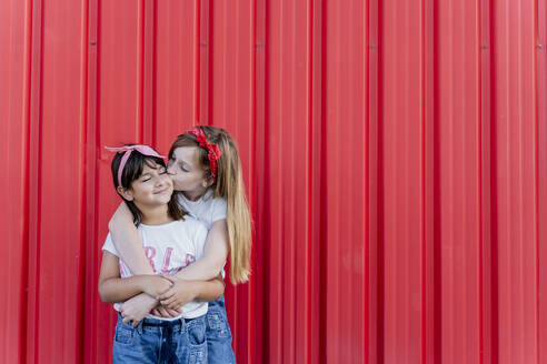 Two sisters standing in front of a red wall, kissing - ERRF01616