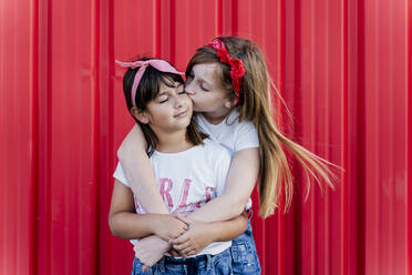 Two sisters standing in front of a red wall, kissing - ERRF01617