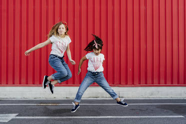 Sisters jumping on street in front of red wall - ERRF01624