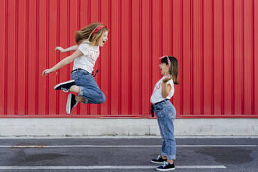 Sisters jumping on street in front of red wall - ERRF01626