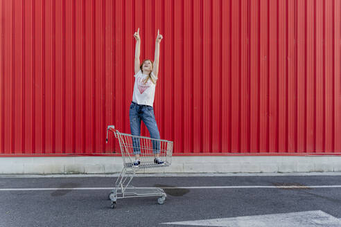 Girl in a shopping cart pointing fingers up in front of red wall - ERRF01647