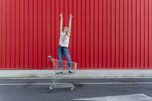 Mädchen in einem Einkaufswagen zeigt mit den Fingern nach oben vor einer roten Wand - ERRF01647