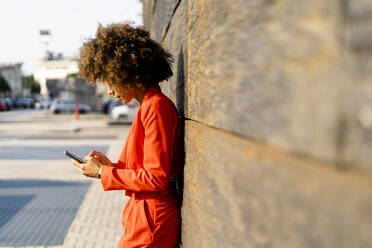 Junge Frau in modischem rotem Hosenanzug lehnt an der Wand und benutzt ein Mobiltelefon - GIOF06864