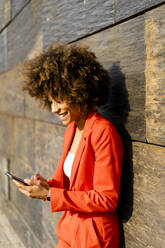 Happy young woman wearing fashionable red pantsuit leaning against wall looking at cell phone - GIOF06865