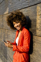 Portrait of happy young woman with smartphone wearing fashionable red pantsuit leaning against wall - GIOF06867