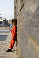 Smiling young woman with cell phone wearing fashionable red pantsuit leaning against wall - GIOF06869