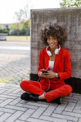 Porträt einer lächelnden jungen Frau mit Smartphone und Kopfhörern in einem modischen roten Hosenanzug - GIOF06875