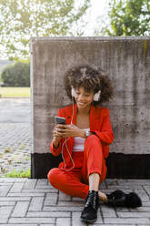 Portrait of smiling young woman with smartphone and headphones wearing fashionable red pantsuit - GIOF06878