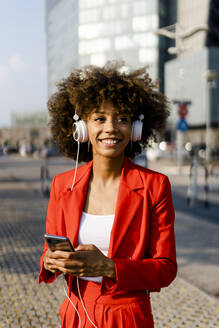 Porträt einer lächelnden jungen Frau mit Smartphone und Kopfhörern in einem modischen roten Hosenanzug - GIOF06882