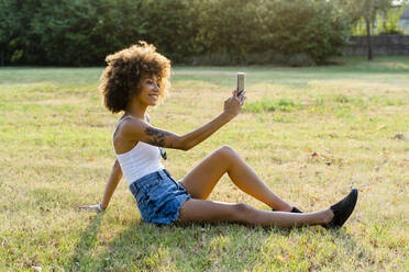 Tätowierte junge Frau sitzt auf einer Wiese und macht ein Selfie mit Smartphone - GIOF06912