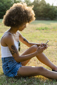 Tätowierte junge Frau sitzt im Sommer auf einer Wiese und benutzt ihr Smartphone - GIOF06914