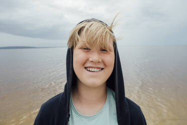 Portrait of happy blond boy with freckles wearing hooded jacket in front of Zeya River, Russia - VPIF01389