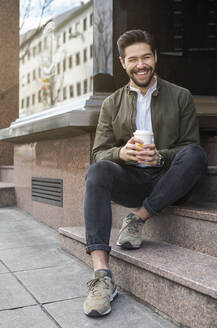 Portrait of laughing young man sitting on steps with coffee to go - JUNF01680